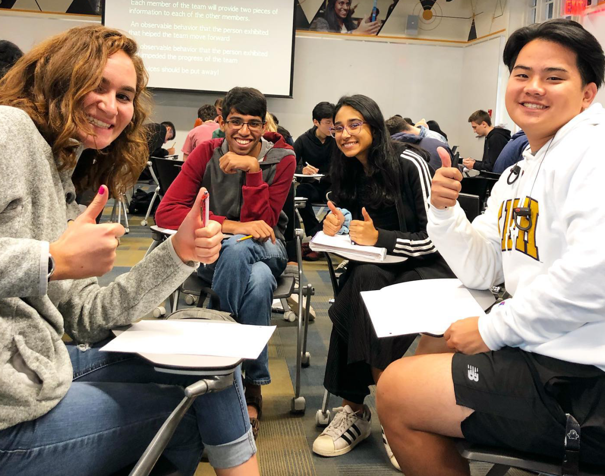 Four smiling students sitting down in a classroom setting: One blond, white female, two brown skinned (1 female-1 male) and one Asian male.
